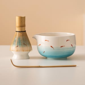 May include: A matcha tea set comprising a white ceramic bowl with a blue watercolour design and red fish, a bamboo whisk, and a bamboo scoop. The whisk has a light blue and tan colour scheme.