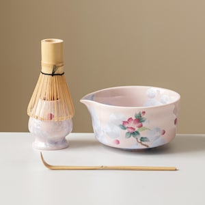 May include: A matcha tea set comprising a light pink ceramic bowl with a floral pattern, a bamboo whisk, and a bamboo scoop. The bowl has a spout. The whisk is made of thin bamboo bristles.