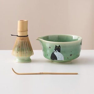 以下が含まれることがあります： 黒と白の猫のデザインが施された緑色のセラミック製の抹茶ボウル、竹製の抹茶ホイスク、竹製の抹茶スプーン