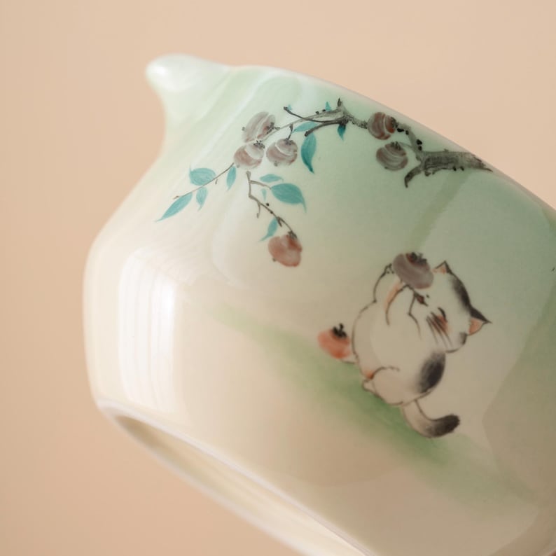 May include: A light green ceramic teacup with a spout and a painted illustration of a cat and a branch with fruit. The teacup has a glossy finish and a curved shape.