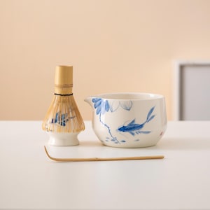Puede incluir: Una taza de té de cerámica blanca con un diseño floral azul y blanco y un pez. También se incluye un batidor de bambú y una cuchara de té de bambú.