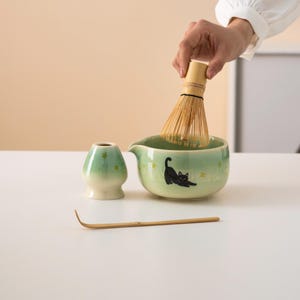 Pu&ograve; includere: Una ciotola di matcha in ceramica verde con un design di gatto nero, una frusta di bamb&ugrave;, una tazza da t&egrave; in ceramica verde abbinata e uno scoop di matcha in bamb&ugrave; su una superficie bianca.