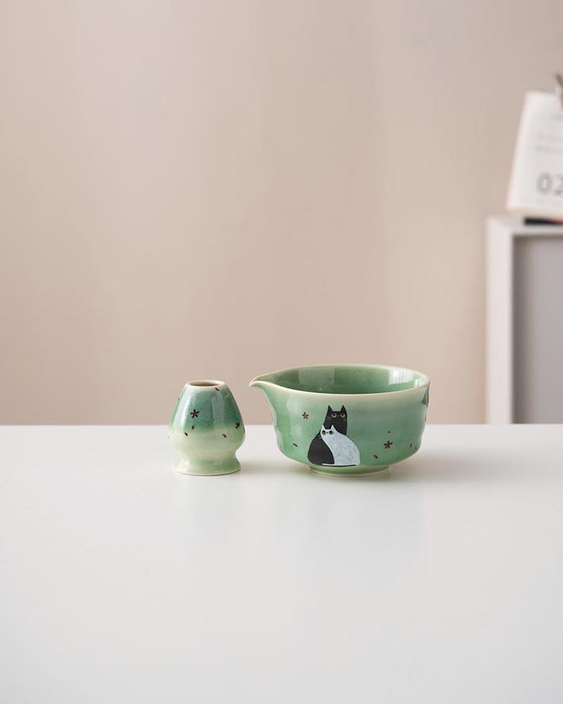 May include: A green ceramic teacup and a matching tea whisk with a black and white cat design. The teacup has a small spout and the whisk has a rounded base.
