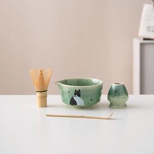 May include: A green ceramic matcha bowl with a black and white cat design, a bamboo matcha whisk, and a green ceramic matcha teacup.