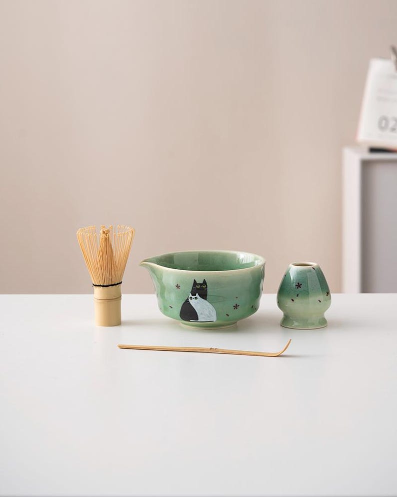 May include: A green ceramic matcha bowl with a black and white cat design, a bamboo matcha whisk, and a green ceramic matcha teacup.