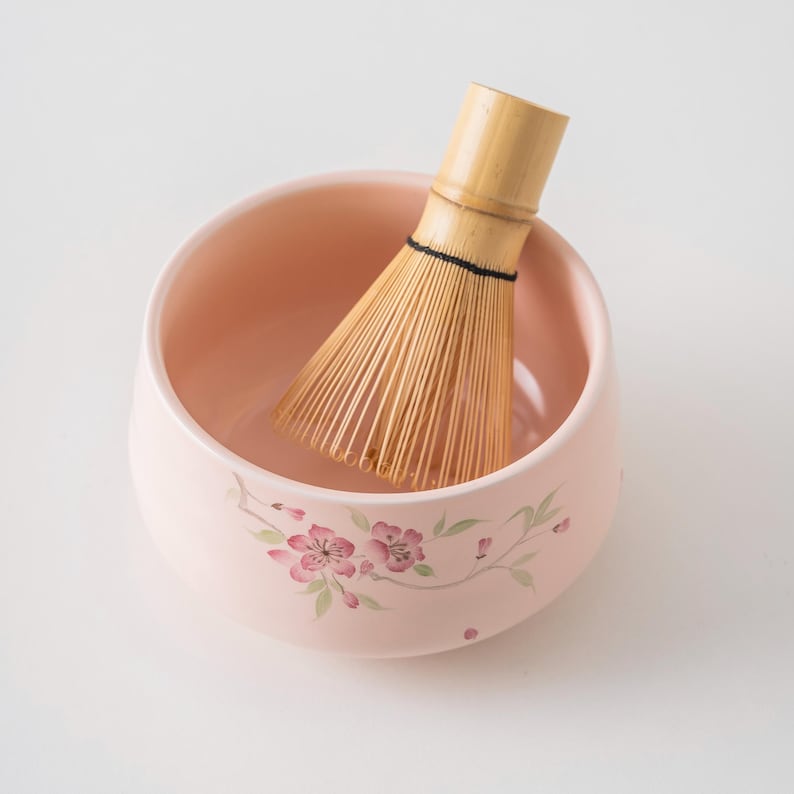 May include: A light pink ceramic matcha bowl with a bamboo whisk inside. The bowl is decorated with pink floral designs. The whisk is made of thin bamboo tines bound together with a black thread.