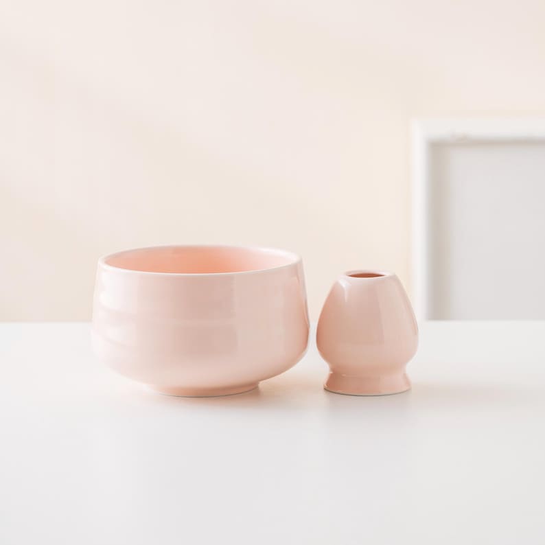May include: A light pink ceramic matcha bowl and whisk holder set. The bowl is wide and shallow, while the whisk holder is small and rounded. Both items are on a white surface, ideal for a tea ceremony.