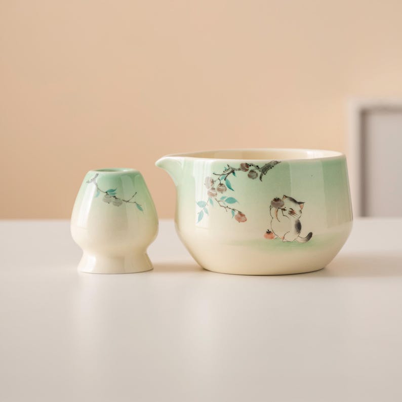 May include: A ceramic tea set featuring a small, rounded matcha bowl and a larger pitcher with a spout. Both pieces are cream-colored with a light green gradient and floral and cat illustrations.