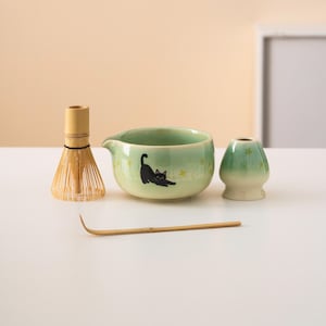 Pu&ograve; includere: Una ciotola di matcha in ceramica verde con un design di gatto nero, una frusta di bamb&ugrave;, una paletta di matcha di bamb&ugrave; e una tazza di matcha in ceramica verde.