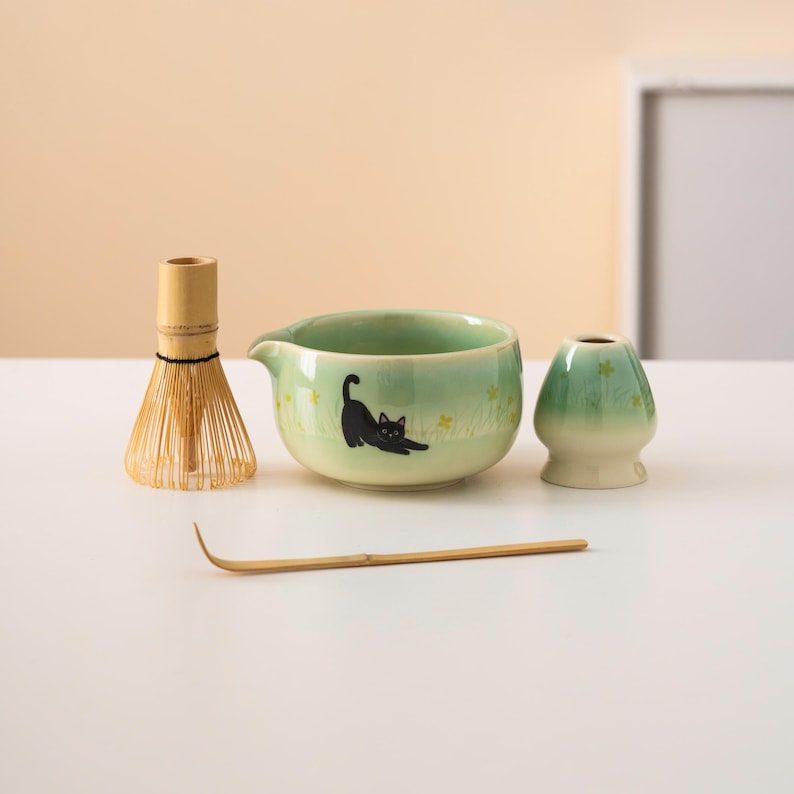 Pu&ograve; includere: Una ciotola di matcha in ceramica verde con un design di gatto nero, una frusta di bamb&ugrave;, una paletta di matcha di bamb&ugrave; e una tazza di matcha in ceramica verde.