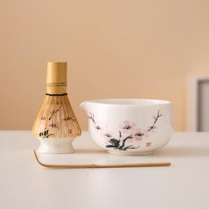 Può includere: Una ciotola di matcha in ceramica bianca con un design floreale rosa e nero, una frusta di matcha in bambù e una paletta di matcha in bambù su una superficie bianca.