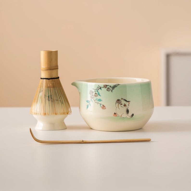 May include: A matcha tea set featuring a bamboo whisk, a ceramic bowl with a cat illustration, and a bamboo scoop. The bowl has a light green gradient and floral accents. The whisk is made of natural bamboo.