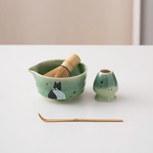 May include: A green ceramic matcha bowl with a black and white cat design, a bamboo whisk, a green ceramic matcha cup, and a bamboo matcha scoop.