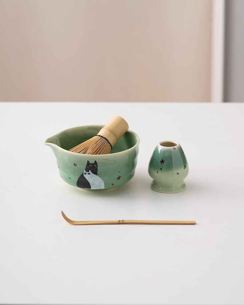 May include: A green ceramic matcha bowl with a black and white cat design, a bamboo whisk, a green ceramic matcha cup, and a bamboo matcha scoop.