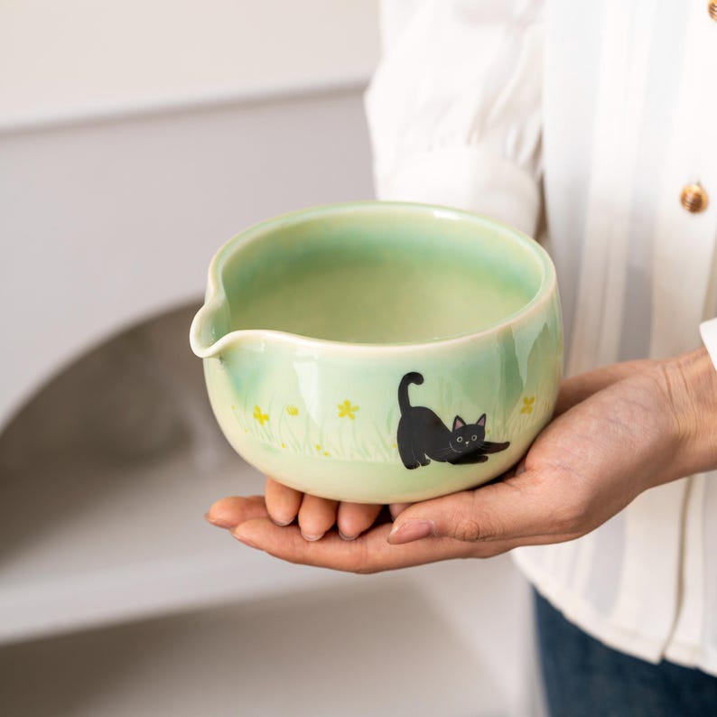 Pu&ograve; includere: Una ciotola in ceramica verde con un design di gatto nero. Il gatto cammina attraverso un campo di fiori gialli. La ciotola ha un beccuccio per versare.