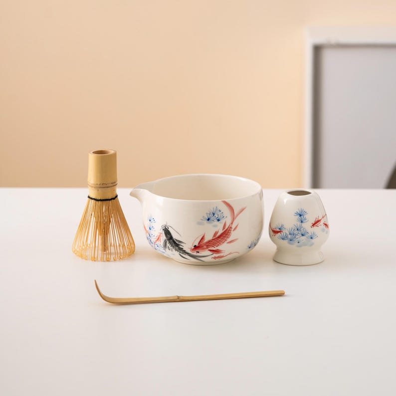 Pu&ograve; includere: Una ciotola di matcha in ceramica bianca con un design di pesce nero e rosso, una frusta di matcha in bamb&ugrave;, una paletta di matcha in bamb&ugrave; e un supporto per frusta di matcha in bamb&ugrave;.