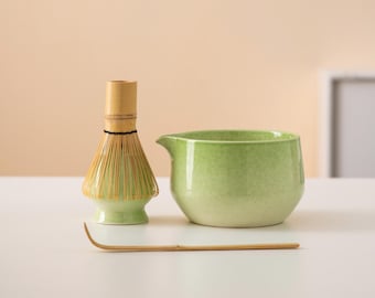 May include: A green ceramic matcha bowl with a bamboo whisk and a wooden spoon on a white surface.