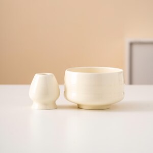 May include: A cream-coloured ceramic matcha bowl and whisk holder sit on a white surface. The bowl is wide and shallow, while the whisk holder is small and egg-shaped. The background is a neutral beige.