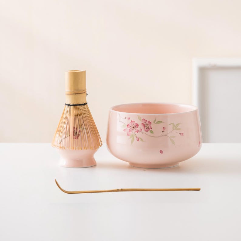 May include: A light pink ceramic matcha bowl and whisk set. The bowl features a floral design in pink and green. The whisk is made of bamboo and has a light pink base. A bamboo scoop is also included.