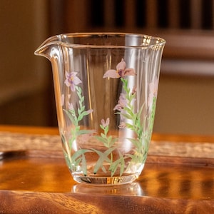 May include: A clear glass pitcher with a floral design. The pitcher has a wide mouth and a handle. The flowers are purple and green and are painted on the glass.
