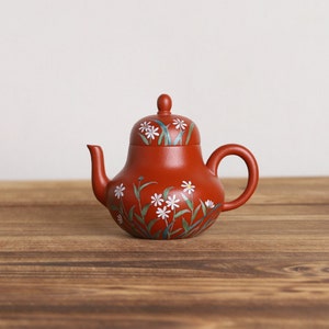 May include: A red clay teapot with a floral design. The teapot has a round body and a lid with a knob. The floral design features white flowers and green leaves.