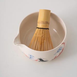 Pu&ograve; includere: Una ciotola di matcha in ceramica bianca con un motivo floreale e una frusta di bamb&ugrave; all'interno. La ciotola ha un beccuccio sul lato.