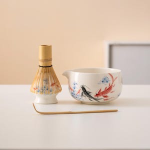 May include: A white ceramic matcha bowl with a blue and red fish design, a bamboo matcha whisk, and a bamboo matcha spoon.