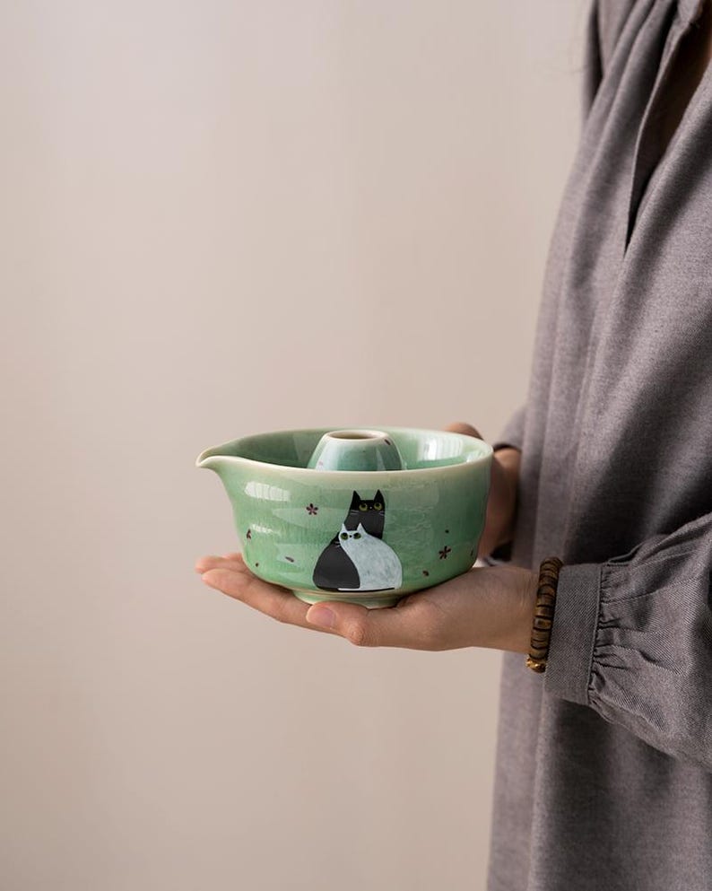 May include: A green ceramic tea infuser with a cat design. The infuser has a small spout and a lid with a hole in the center.