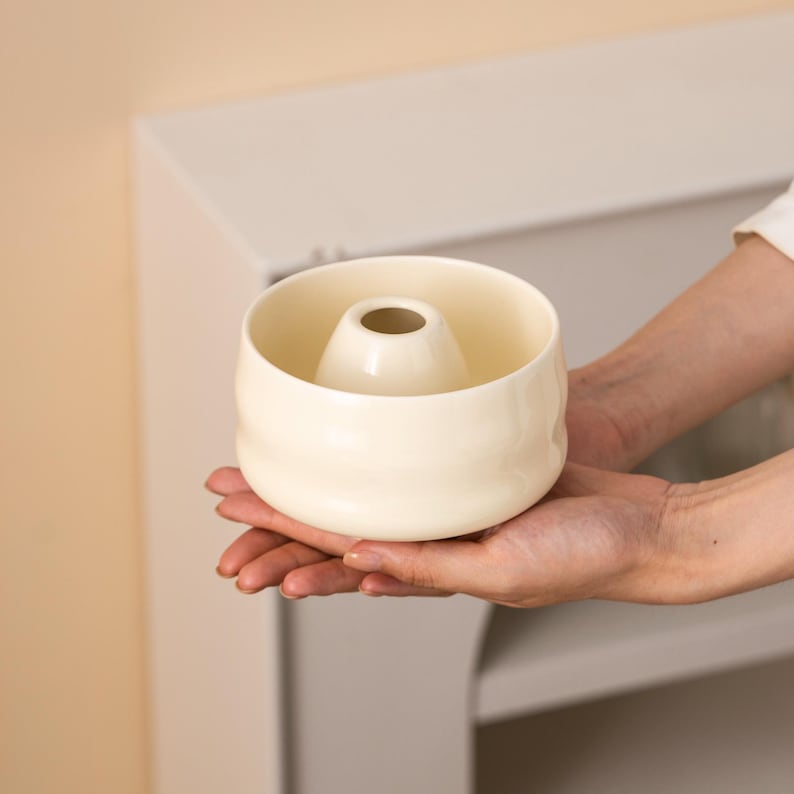 May include: A cream-coloured ceramic bowl with a central, raised cylinder. The bowl is held in open hands, showcasing its smooth, rounded form. The design is simple and elegant, suggesting a minimalist aesthetic.