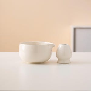 Pu&ograve; includere: Un'anfora in ceramica bianca e un piccolo vaso in ceramica bianca su una superficie bianca.