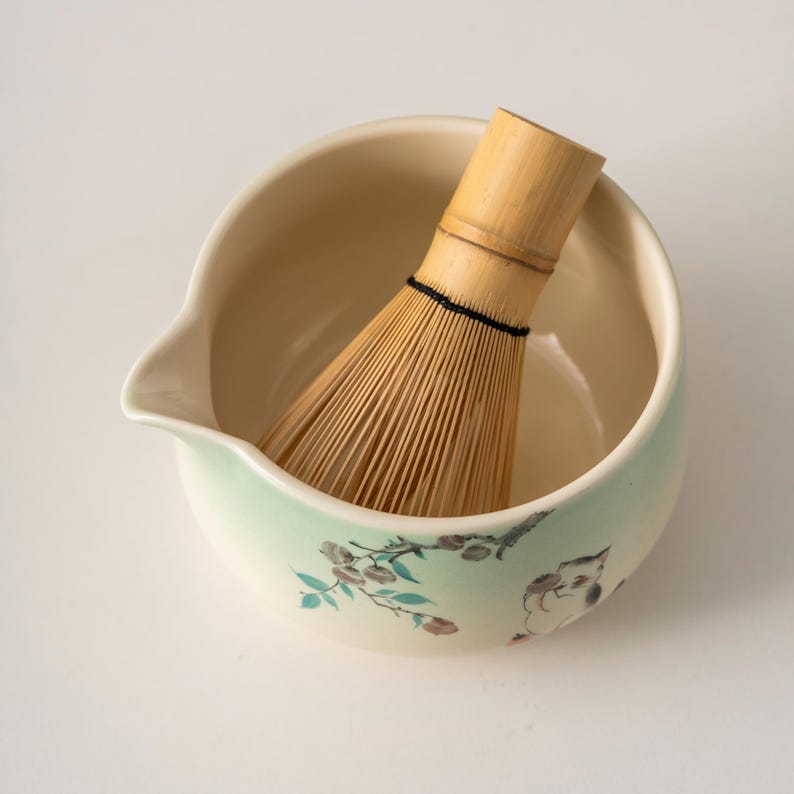 May include: A matcha bowl with a bamboo whisk. The ceramic bowl is off-white with a light green rim and floral design. The whisk is made of bamboo and is inside the bowl. This set is used for preparing matcha tea.