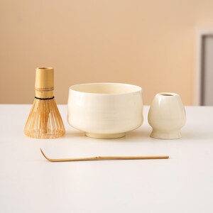 May include: A matcha tea set comprising a bamboo whisk, a ceramic bowl, a small ceramic container, and a bamboo scoop. The bowl and container are off-white, and the whisk and scoop are made of natural bamboo.