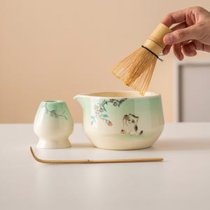 May include: A matcha tea set with a light green and cream-colored ceramic bowl, whisk, and holder. The bowl features a hand-painted cat and floral design. A bamboo whisk is held above the bowl, ready for use. A bamboo scoop rests in front.