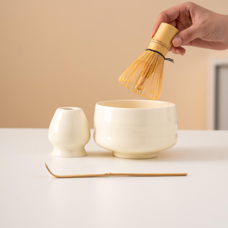 May include: A cream-coloured matcha tea set, including a bamboo whisk, a ceramic bowl, a small ceramic holder, and a bamboo scoop. The whisk is held above the bowl. The set is on a white surface.