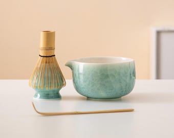Puede incluir: Un tazón de matcha de cerámica verde con un batidor de bambú y una cuchara de matcha de madera sobre una superficie blanca.