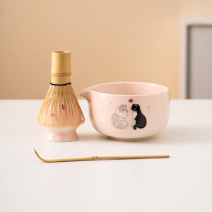 Könnte beinhalten: Ein Matcha-Teeset mit einer rosa Keramikschale mit Katzenillustrationen, einem Bambusbesen und einem Bambuslöffel. Die Schale hat einen Ausguss und ein Blumendesign. Der Schneebesen ist hellbraun und der Löffel ist dünn und gebogen.