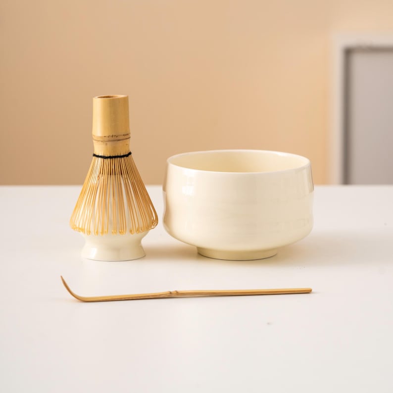 May include: A matcha tea set comprising a cream-coloured ceramic bowl, a bamboo whisk, and a bamboo scoop. The whisk has a light-coloured handle and a fan of thin tines. The bowl has a rounded shape. The scoop is curved.