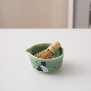 May include: A green ceramic matcha bowl with a black and white cat design and a bamboo whisk resting inside.