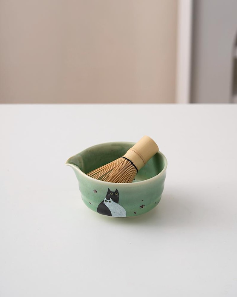 May include: A green ceramic matcha bowl with a black and white cat design and a bamboo whisk resting inside.