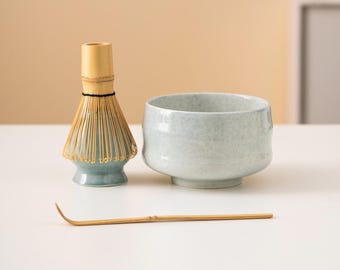 May include: A matcha tea set featuring a light blue ceramic bowl, a bamboo whisk, and a bamboo scoop. The whisk has a light blue base and a natural bamboo handle. The bowl has a speckled appearance.
