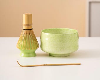May include: A matcha tea set featuring a green ceramic bowl, a bamboo whisk, and a bamboo scoop. The bowl and whisk base are a light green color. The whisk has many bamboo tines. The scoop is a light brown color.