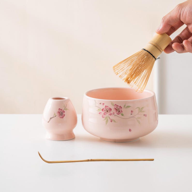 May include: A pink ceramic matcha bowl set with floral designs. The set includes a bowl, a small vase, a bamboo whisk, and a bamboo scoop. The whisk is held by a hand. The bowl and vase have pink cherry blossom designs.