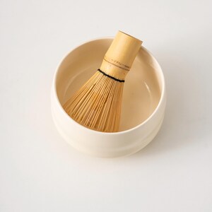 May include: A cream-coloured ceramic matcha bowl containing a bamboo whisk. The whisk is made of thin, light-brown bamboo tines bound with black thread. The bowl has a rounded shape and a smooth surface, ideal for preparing matcha tea.