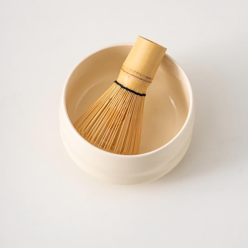 May include: A cream-coloured ceramic matcha bowl containing a bamboo whisk. The whisk is made of thin, light-brown bamboo tines bound with black thread. The bowl has a rounded shape and a smooth surface, ideal for preparing matcha tea.