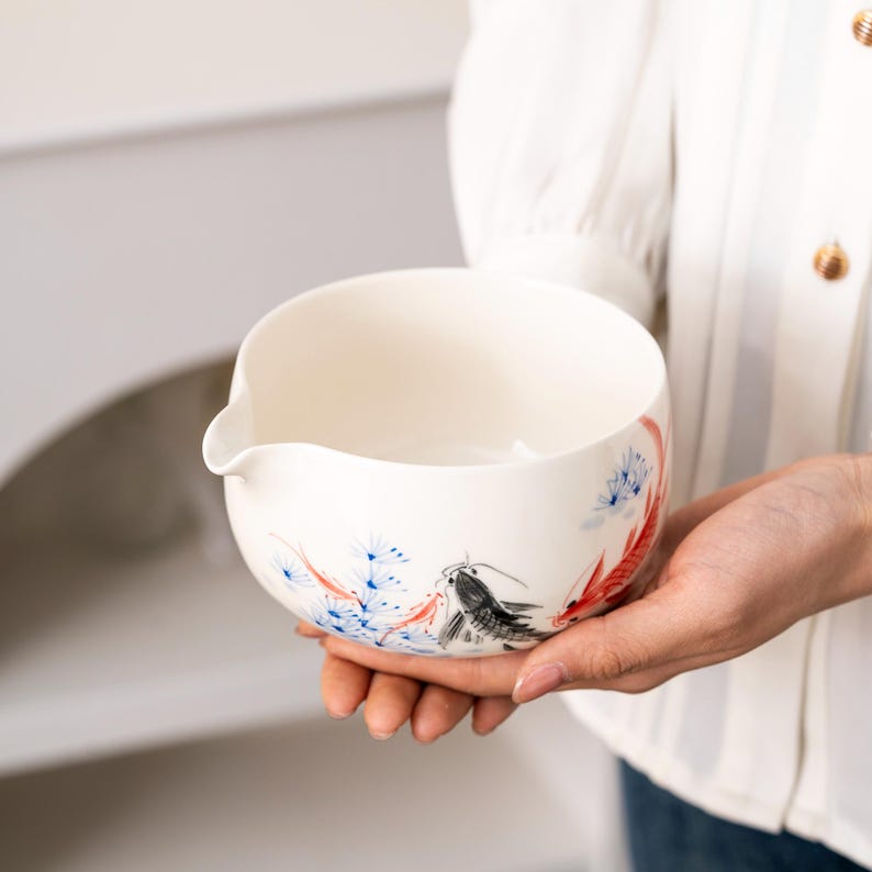 Pu&ograve; includere: Scodella in ceramica bianca con un design floreale blu e rosso e un design di pesce nero e rosso. La ciotola ha un beccuccio su un lato.