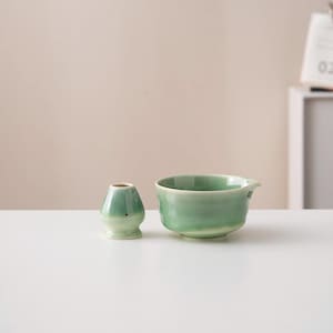 May include: A green ceramic tea bowl and a matching green ceramic tea whisk holder. The bowl has a slightly flared lip and a rounded bottom. The whisk holder is shaped like a small, rounded vase.