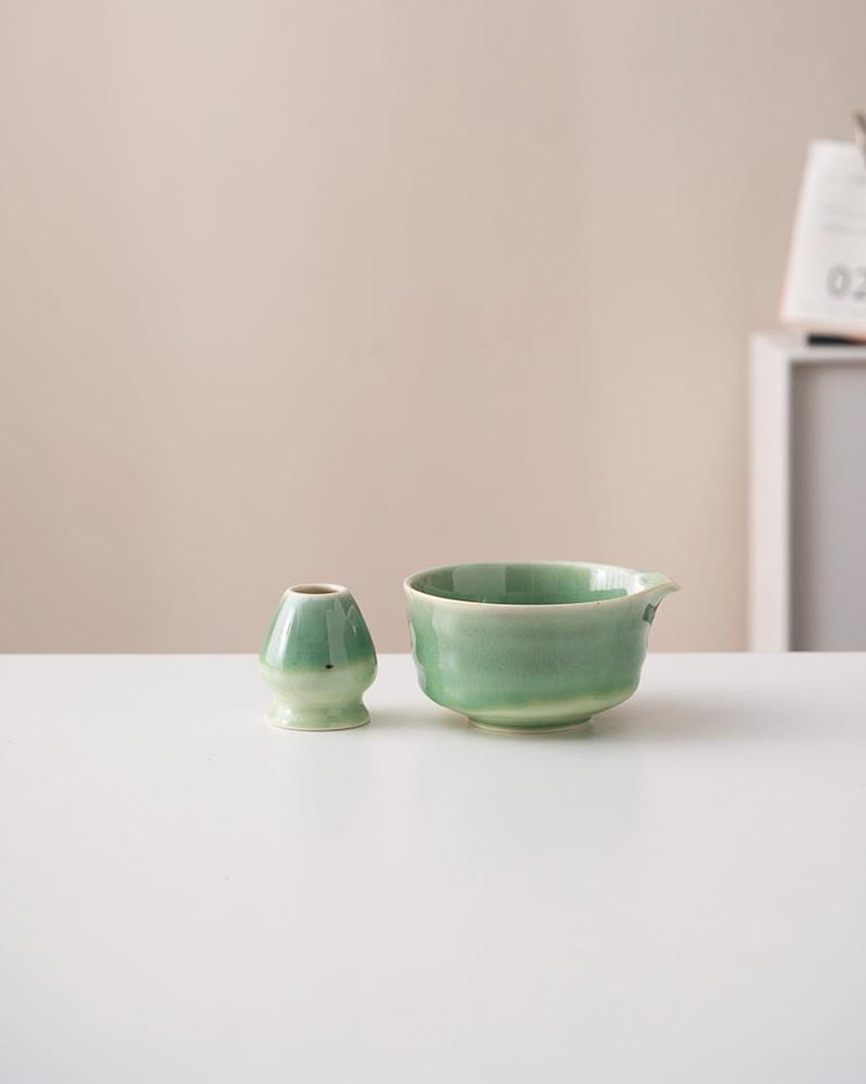 May include: A green ceramic tea bowl and a matching green ceramic tea whisk holder. The bowl has a slightly flared lip and a rounded bottom. The whisk holder is shaped like a small, rounded vase.
