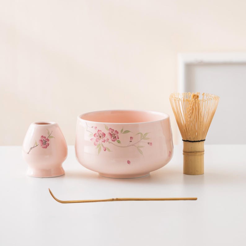 May include: A pink ceramic matcha tea set with floral designs. The set includes a bowl, a whisk, a small vase, and a bamboo scoop. The bowl and vase are decorated with pink cherry blossoms and green leaves.