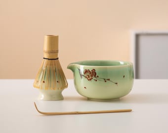 May include: A green ceramic matcha bowl with a floral design, a bamboo matcha whisk, and a bamboo whisk holder on a white surface.