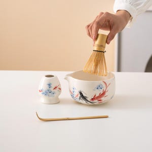 Pu&ograve; includere: Una ciotola di matcha in ceramica bianca con un design di pesci blu e rossi, un porta-frusta per matcha in ceramica bianca con un design floreale blu e rosso, una frusta per matcha in bamb&ugrave; e una paletta per matcha in bamb&ugrave;.